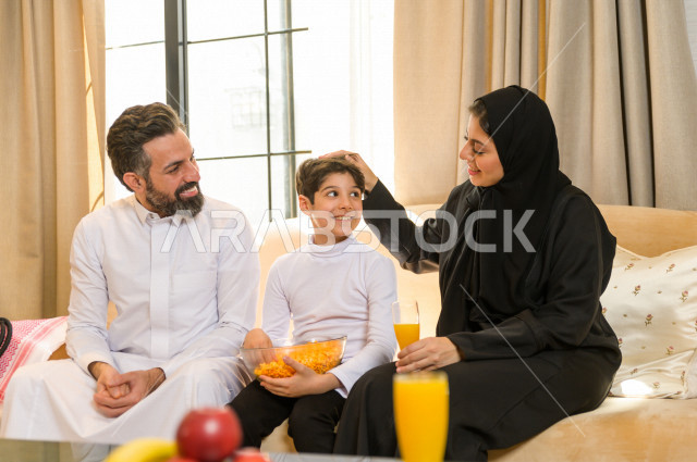 أسرة خليجية صغيرة وسعيدة تجلس على الأريكة في المنزل ، الاستمتاع بقضاء الوقت مع العائلة ، يوم الطفل العالمي ، عائلة سعودية ، اجواء عائلية سعيدة 