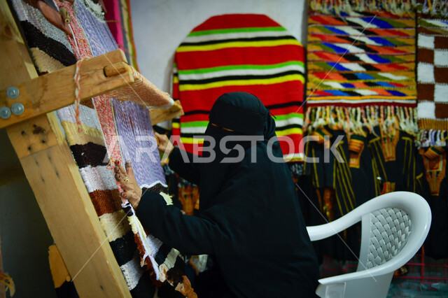 صناعات ومهن تقليدية شعبية قديمة، حرف يدوية تراثية سعودية، صورة من الخلف لإمرأة عربية خليجية سعودية ترتدي العباءة السوداء تقوم بحياكة وغزل النسيج والأقمشة, حرفة صناعة المشالح والمفارش، استخدام الخيوط الملونة، الفنون والمهارات النسائية