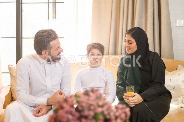 أسرة خليجية صغيرة وسعيدة تجلس على الأريكة في المنزل ، الاستمتاع بقضاء الوقت مع العائلة ، يوم الطفل العالمي ، عائلة سعودية ، اجواء عائلية سعيدة 
