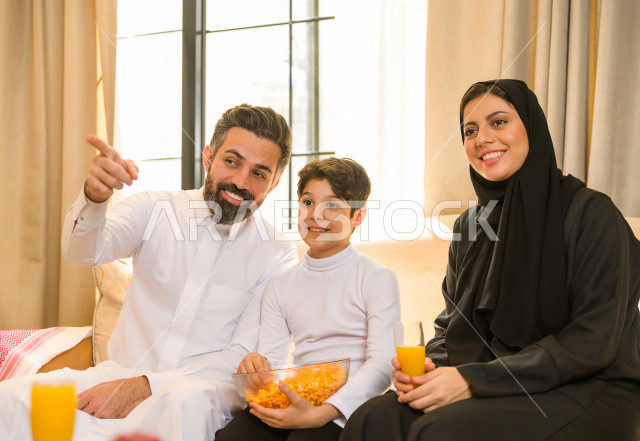 أسرة خليجية صغيرة وسعيدة تجلس على الأريكة في المنزل ، الاستمتاع بقضاء الوقت مع العائلة ، يوم الطفل العالمي ، عائلة سعودية ، اجواء عائلية سعيدة 