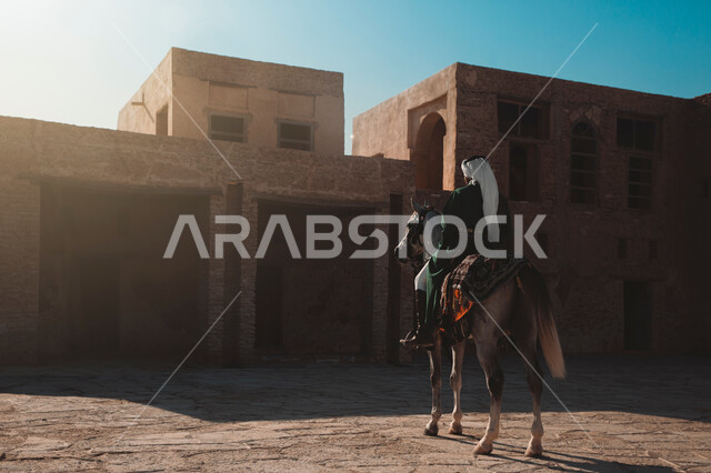 التجول في المناطق التاريخية الأثرية، صورة مقربة من الخلف لرجل عربي خليجي سعودي يرتدي الثوب التقليدي يمتطي حصانه يقف بجانب بيت طيني، الأماكن الأثرية في المملكة العربية السعودية