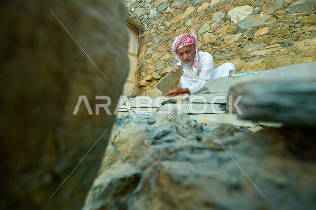 إعادة بناء وترميم البيوت والمنشآت القديمة, مفهوم التمسك بالأصالة والتراث السعودي, رجل كبير بالسن عربي خليجي سعودي يرتدي الثوب التقليدي والشماغ يمسك بيده الحجارة, الآثار والأماكن التاريخية الشعبية