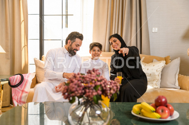 أسرة خليجية صغيرة وسعيدة تجلس على الأريكة في المنزل ، الاستمتاع بقضاء الوقت مع العائلة ، يوم الطفل العالمي ، عائلة سعودية ، اجواء عائلية سعيدة 