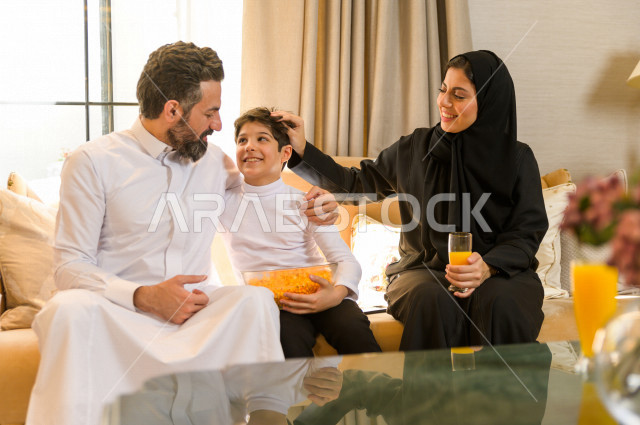 أسرة خليجية صغيرة وسعيدة تجلس على الأريكة في المنزل ، الاستمتاع بقضاء الوقت مع العائلة ، يوم الطفل العالمي ، عائلة سعودية ، اجواء عائلية سعيدة 