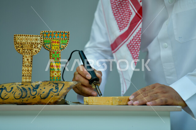 مهن وحرف شعبيه تراثية ،الأصالة والعراقة والفخر و الاعتزاز بالتراث السعودي، الرسم والتخطيط اليدوي، صورة مقربة لأيدي رجل عربي خليجي سعودي يرتدي الزي التقليدي يقوم بالنقش على قطع خشبية، تحف أثرية تقليدية