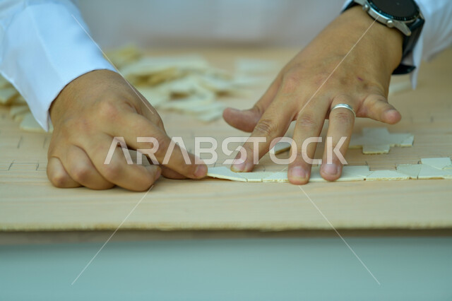 Popular heritage works, Saudi professions and jobs, intelligence and concentration games, the concept of puzzle solving, amusement and entertainment, a close-up picture of the hands of a Saudi Gulf Arab man wearing a smart watch, standing in one of the shops and cutting the puzzle pieces.