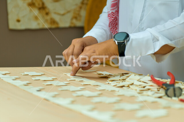 مهن ووظائف سعودية ،ألعاب الذكاء و التركيز ،مفهوم حل اللغز و التسلية والترفيه، صورة مقربة لأيدي رجل عربي خليجي سعودي يرتدي الساعه الذكية و يقف في أحد المحلات التجارية ويقوم بقص قطع البازل، أعمال تراثية شعبية