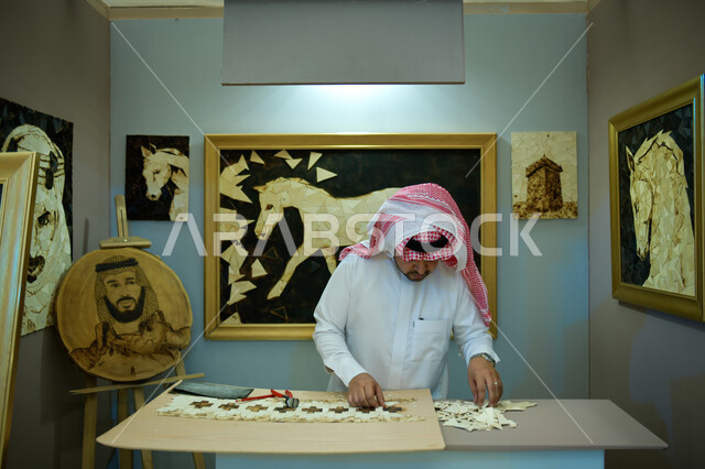 لوحات فنية معلقة، مفهوم التركيز وحل اللغز و التسلية والترفيه، رجل عربي خليجي سعودي يرتدي الزي التقليدي يقف في أحد المحلات التجارية ويقوم بقص قطع البازل، أعمال تراثية شعبية، مهن ووظائف سعودية