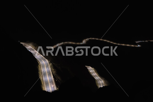 طريق اسفلتي ملتوي وعر وسط الجبال الصخرية في منطقة عقبة الهدا في الليل، القمم والمرتفعات الجبلية في المملكة العربية السعودية، صورة جوية من الأعلى لحركة مرور وسير السيارات على جبال الهدا في مدينة الطائف