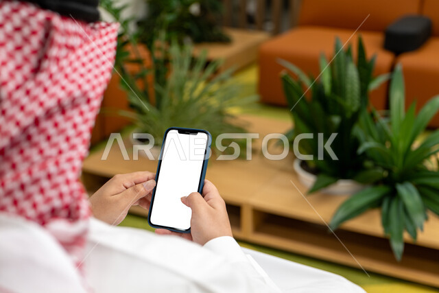 Using modern and advanced technology devices, displaying a blank white screen on a mobile phone, a close-up picture of the hand of a Saudi Gulf Arab man wearing a traditional thobe and shemagh, holding a mobile phone, enjoying quiet sessions.