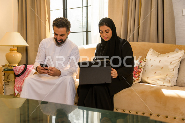 زوجان خليجيان سعيدان يجلسان على الأريكة في المنزل ، يستخدمان الابتوب المحمول ، الاستمتاع بقضاء الوقت في المنزل ، قضاء وقت الفراغ مع العائلة ، اسره سعودية خليجية