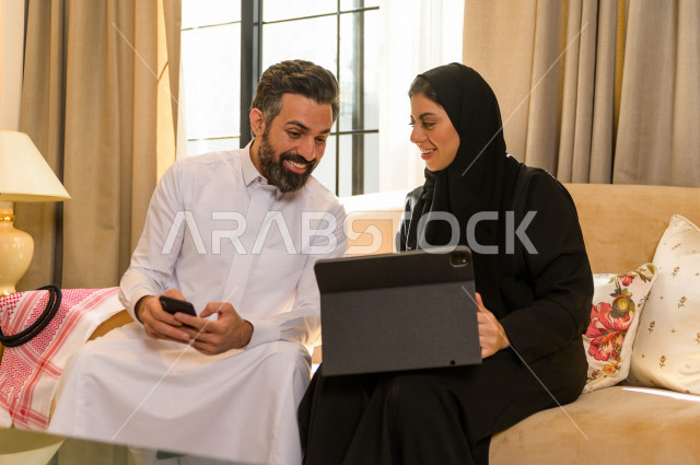 زوجان خليجيان سعيدان يجلسان على الأريكة في المنزل ، يستخدمان الابتوب المحمول ، الاستمتاع بقضاء الوقت في المنزل ، قضاء وقت الفراغ مع العائلة ، اسره سعودية خليجية