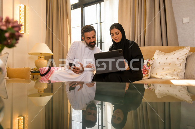 زوجان خليجيان سعيدان يجلسان على الأريكة في المنزل ، يستخدمان الابتوب المحمول ، الاستمتاع بقضاء الوقت في المنزل ، قضاء وقت الفراغ مع العائلة ، اسره سعودية خليجية