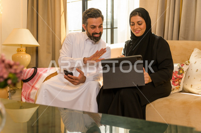 زوجان خليجيان سعيدان يجلسان على الأريكة في المنزل ، يستخدمان الابتوب المحمول ، الاستمتاع بقضاء الوقت في المنزل ، قضاء وقت الفراغ مع العائلة ، اسره سعودية خليجية