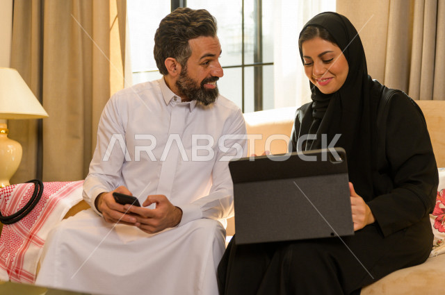 زوجان خليجيان سعيدان يجلسان على الأريكة في المنزل ، يستخدمان الابتوب المحمول ، الاستمتاع بقضاء الوقت في المنزل ، قضاء وقت الفراغ مع العائلة ، اسره سعودية خليجية