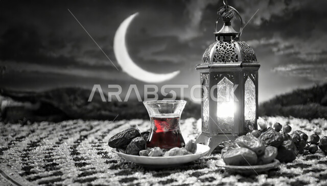 الاستعداد لاستقبال شهر الخير والبركة، هلال شهر رمضان المبارك ,أعياد ومناسبات دينية ،صورة بالابيض والاسود لفانوس معدني تقليدي مضيء مع طبق من حبات التمر الطازجة وكأس من الشاي ،أجواء رمضانية روحانية
