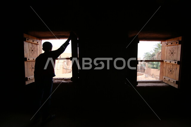 الآثار التاريخية العريقة في المملكة العربية السعودية، الأماكن و المعالم السياحية المشهورة، صورة من الخلف لرجل عربي خليجي سعودي يقف في داخل احدى البيوت الطينية القديمة أمام نافذة خشبية مطلة على أحياء قرية أشيقر في مدينة الرياض