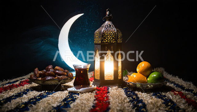 أجواء رمضانية روحانية، الاستعداد لقدوم شهر الخير والبركة، فانوس معدني تقليدي مضيء مع اطباق متنوعه من الاغذية الصحية و حبات التمر الطازجة وكوب من الشاي، أعياد ومناسبات دينية إسلامية، خلفية هلال شهر رمضان في السماء