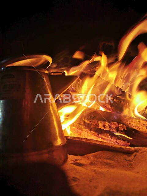 جلسة بر عربية شعبية، صورة مقربة لدلة قهوة معدنية أمام شعلة من الحطب في الصحراءد، النيران المشتعلة على الرمال الذهبية الناعمة، المناطق الصحراوية في المملكة العربية السعودية، الاستمتاع بالأجواء الشتوية والمشروبات الساخنة