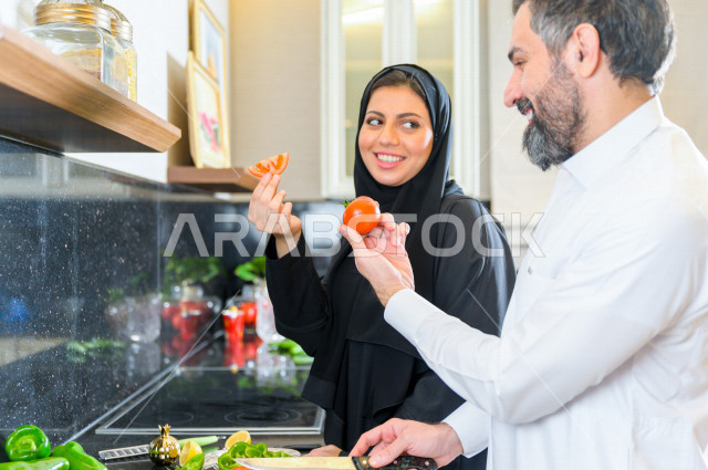 زوجان سعوديان خليجيان يتشاركان في اعداد وجبه الطعام بالمطبخ ، الاستمتاع باداء الاعمال المنزلية ، ايماءات و تعابير وجه تدل على الفرحة