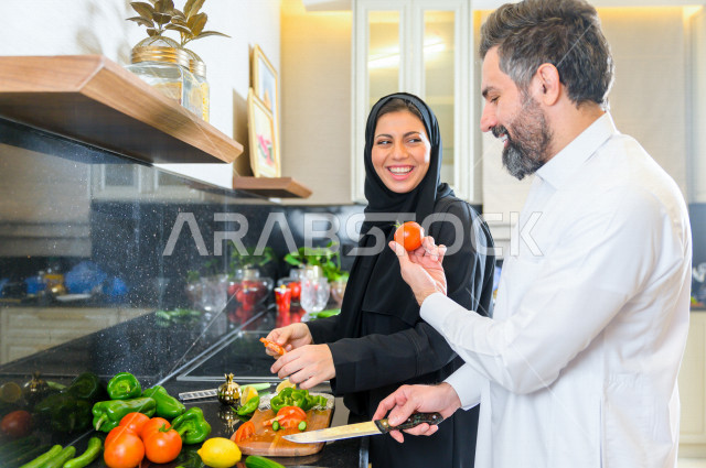 زوجان سعوديان خليجيان يتشاركان في اعداد وجبه الطعام بالمطبخ ، الاستمتاع باداء الاعمال المنزلية ، ايماءات و تعابير وجه تدل على الفرحة