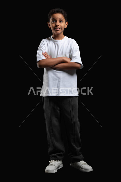 الوقوف بإيماءات أيدي متقاطعة، بورتريه لفتى عربي سعودي خليجي يرتدي الزي الكاجوال ينظر الى الكاميرا بتعبيرات الدهشة، الذهاب الى السينما لقضاء وقت ممتع، مشاهدة أفلام الكرتون على الشاشة الكبيرة، صورة الجسم بالطول الكامل، خلفية سوداء