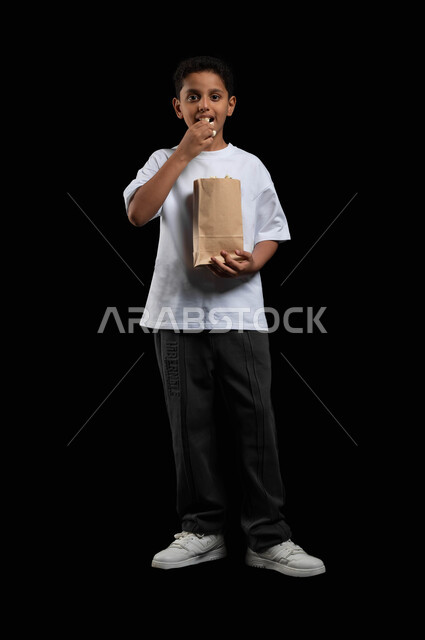 ايماءات السعادة للذهاب الى السينما، الاستمتاع بتجربة سينمائية مثيرة، تذوق طعم البوبكورن الطازج والمقرمش خلال مشاهدة الفلم، بورتريه لفتى عربي خليجي سعودي مبتسم يرتدي الملابس الكاجوال ينظر الى الكاميرا بابتسامة، صورة الجسم بالطول الكامل، خلفية سوداء