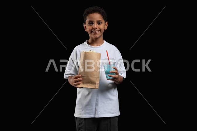 تناول العصائر المنعشة في السينما، ايماءات السعادة والاستمتاع بمتابعة الفلم، الترفيه عن النفس وقضاء وقت ممتع، بورتريه لفتى عربي خليجي مبتسم يرتدي الملابس الكاجوال ويمسك بيده كأس من عصير الموهيتو الشهي وكيس ورقي من الفوشار، خلفية سوداء