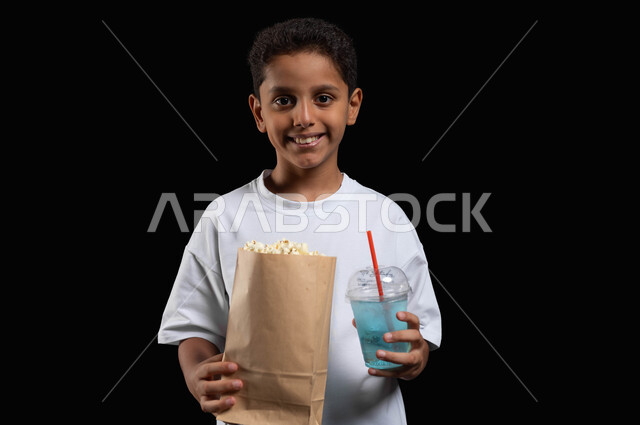 قضاء أوقات ممتعة في العطلة، الاستمتاع بالذهاب الى السينما، تناول عصير الموهيتو المنعش أثناء مشاهدة الفلم،  بورتريه مقرب لفتى عربي خليجي مبتسم يرتدي الملابس الكاجوال ويمسك بيده كأس من المشروب البارد وكيس ورقي من الفوشار، خلفية سوداء