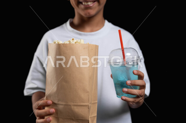 الاستمتاع بالذهاب الى السينما، تناول عصير الموهيتو المنعش أثناء مشاهدة الفلم، قضاء أوقات ممتعة في العطلة، بورتريه مقرب لفتى عربي خليجي مبتسم يرتدي الملابس الكاجوال ويمسك بيده كأس من المشروب البارد وكيس ورقي من الفوشار، خلفية سوداء
