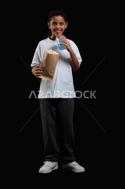 Drinking refreshing mojito juice while watching the movie, enjoying going to the cinema, spending fun times on vacation, portrait of a smiling Gulf Arab boy wearing casual clothes and holding a cup of cold drink in his hand and a paper bag of popcorn, full body portrait, black background