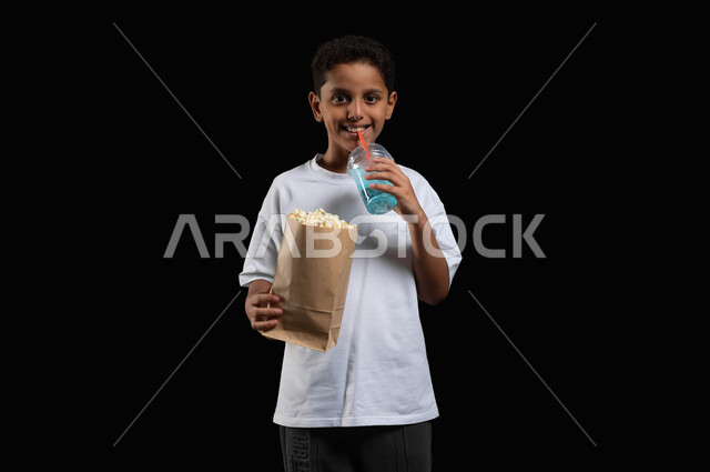 الترفيه عن النفس وقضاء وقت ممتع، ايماءات السعادة والاستمتاع بمتابعة الفلم، تناول المشروبات الباردة في السينما، بورتريه لفتى عربي خليجي مبتسم يرتدي الملابس الكاجوال ويمسك بيده كأس من عصير الموهيتو الشهي وكيس ورقي من الفوشار، خلفية سوداء