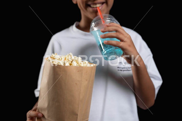 تناول المشروبات الباردة في السينما، ايماءات السعادة والاستمتاع بمتابعة الفلم، الترفيه عن النفس وقضاء وقت ممتع، بورتريه مقرب لفتى عربي خليجي مبتسم يرتدي الملابس الكاجوال ويمسك بيده كأس من عصير الموهيتو الشهي وكيس ورقي من الفوشار، خلفية سوداء