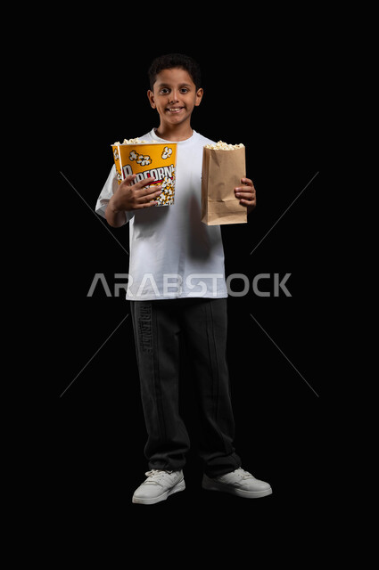 قضاء وقت ممتع في دور السينما، ايماءات الفرح والاستمتاع بمتابعة الفلم، تناول البوبكورن الطازج والمقرمش، بورتريه لفتى عربي سعودي خليجي يرتدي الملابس الكاجوال يمسك كيس ورقي ووعاء من الفوشار في يديه وينظر الى الكاميرا بابتسامة، صورة الجسم بالكامل، خلفية سوداء