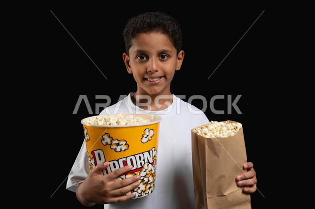 الذهاب الى السينما لقضاء وقت ممتع، تناول البوبكورن الطازج والمقرمش، بورتريه مقرب لفتى عربي سعودي خليجي يرتدي الزي الكاجوال يمسك كيس ورقي ووعاء من الفوشار في يديه وينظر الى الكاميرا بابتسامة، ايماءات الفرح والاستمتاع بمتابعة الفيلم، خلفية سوداء