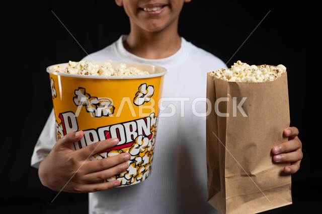 تناول البوبكورن الطازج والمقرمش، الذهاب الى السينما لقضاء وقت ممتع، بورتريه مقرب لفتى عربي سعودي خليجي يرتدي الزي الكاجوال يمسك كيس ورقي ووعاء من الفوشار في يديه بابتسامة، ايماءات الفرح والاستمتاع بمتابعة الفيلم، خلفية سوداء