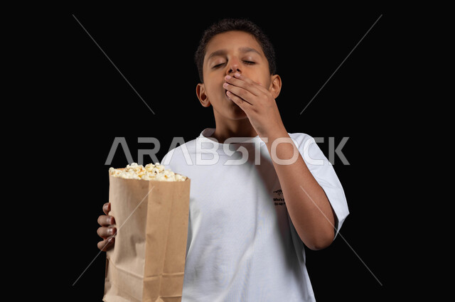 نشاط فردي ترفيهي أثناء الاستراحة في دور السينما، التثاؤب والنعاس والإحساس بالملل خلال مشاهدة الأفلام، بورتريه مقرب لفتى عربي خليجي سعودي يرتدي الزي الكاجوال يغمض عينيه اثناء تناول البوبكورن، خلفية سوداء