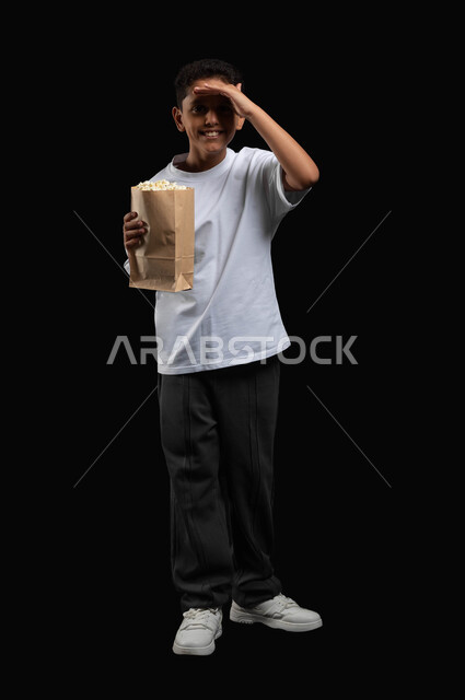 قضاء وقت مسلي وممتع، تناول الفوشار اللذيذ في السينما، متابعة أحداث الفيلم بتركيز واهتمام، بورتريه لفتى عربي خليجي سعودي مبتسم يرتدي الزي الكاجوال يضع يده على رأسه للنظر إلى شيء ما بتمعن، صورة الجسم بالطول الكامل، خلفية سوداء