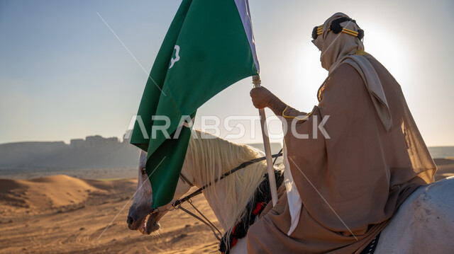  الاحتفال مع الأحصنة في المناسبات الوطنية، يوم بدينا 1727 م، ذكرى تأسيس الدولة السعودية الأولى 22 فبراير، صورة من الخلف لرجل عربي خليجي سعودي يرتدي البشت والغترة يمتطي خيل أبيض أصيل وسط الرمال الذهبية، رفع علم التأسيس في الصحراء، توارث حب الجياد بين أبناء الوطن