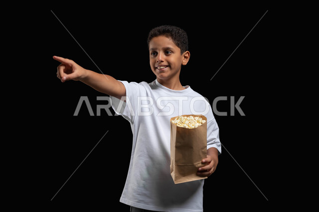 الاستمتاع بأحداث الفيلم أثناء تناول الفوشار، الابتعاد عن ضغوط الحياة اليومية، بورتريه لفتى عربي خليجي سعودي مبتسم يرتدي الملابس الكاجوال يشير باصبعه الى شيء ما، الذهاب الى السينما لقضاء وقت ممتع، خلفية سوداء