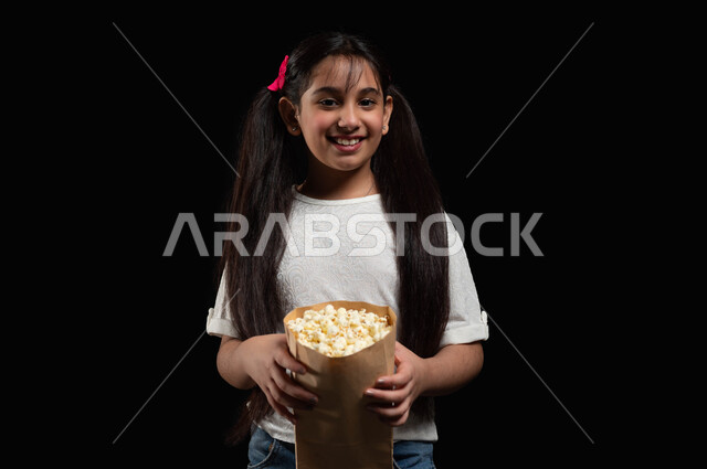 الابتعاد عن روتين الحياة اليومية، بورتريه مقرب لفتاة عربية خليجية سعودية مبتسمة ترتدي الزي الكاجوال تمسك بيدها كيس فوشار ورقي، متابعة الأفلام والعروض المسرحية في السينما، ايماءات الاستمتاع بمشاهدة فيلم ممتع، خلفية سوداء