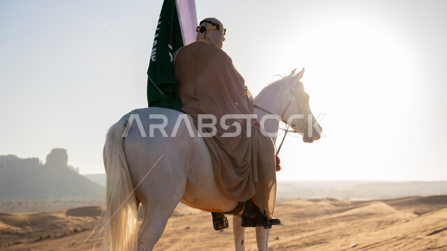 يوم بدينا 1727 م، ذكرى تأسيس الدولة السعودية الأولى 22 فبراير، صورة من الخلف لرجل عربي خليجي سعودي يرتدي البشت والغترة يمتطي خيل أبيض أصيل وسط الرمال الذهبية، رفع علم التأسيس في الصحراء، توارث حب الجياد بين أبناء الوطن، الاحتفال مع الأحصنة في المناسبات الوطنية
