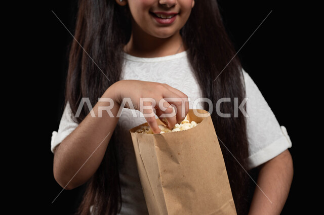 مشاهدة المسلسلات والبرامج التلفزيونية، ايماءات الفرح والسعادة، تناول السناكات والوجبات الخفيفة، بورتريه مقرب لفتاة عربية خليجية سعودية تحمل كيس من الفشار، كسر الروتين والذهاب الى السينما في عطلة نهاية الأسبوع، متعة حضور الأفلام المفضلة، خلفية سوداء