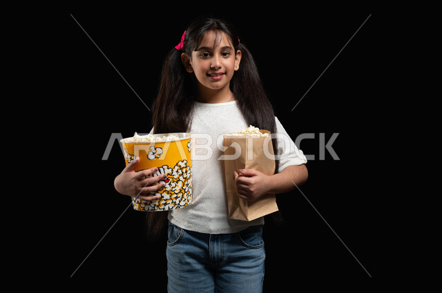 Using your free time to watch favorite movies, getting ready to go to the cinema, watching TV series and programmes, a portrait of a Saudi Arabian Gulf girl holding a lot of popcorn in her hands, gestures of happiness and pleasure, eating snacks and snacks, a black background.