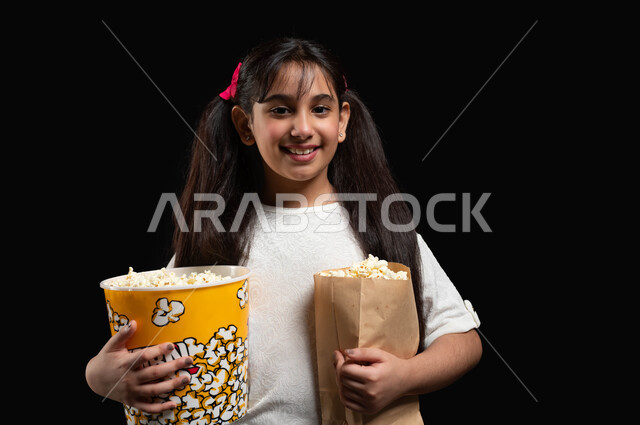 الاستعداد للدخول الى السينما، متابعة المسلسلات والبرامج التلفزيونية، بورتريه مقرب لفتاة عربية خليجية سعودية تحمل بيديها الكثير من الفوشار، استغلال أوقات الفراغ في مشاهدة الأفلام المفضلة، ايماءات السعادة والسرور،تناول الوجبات الخفيفة والسناك، خلفية سوداء