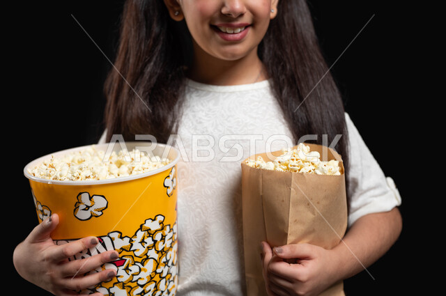 متابعة المسلسلات والبرامج التلفزيونية، الاستعداد للدخول الى السينما، بورتريه مقرب لفتاة عربية خليجية سعودية تحمل بيديها الكثير من الفوشار، استغلال أوقات الفراغ في مشاهدة الأفلام المفضلة، ايماءات السعادة والسرور،تناول الوجبات الخفيفة والسناك، خلفية سوداء