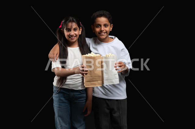 قضاء أوقات الفراغ بمشاهدة البرنامج التلفزيوني المفضل، بورتريه لولد عربي خليجي سعودي يقوم بوضع يده على كتف أخته، الاستمتاع بنكهة الفوشار الطازج، متابعة الأفلام والعروض المسرحية في السينما، النظر الى الكاميرا بايماءات السرور والبهجة، خلفية سوداء