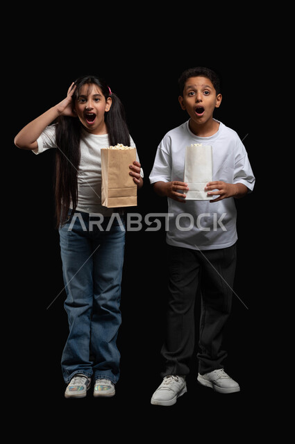 القيام بالأنشطة الترفيهية بالذهاب الى السينما مع الأخوة، قضاء أوقات ممتعة ومسلية في عطلة نهاية الأسبوع، بورتريه لولد وبنت عربيان خليجيان سعوديان يستمتعان بأكل الفوشار ومشاهدة الأفلام، ايماءات وجه ويدين تدل على الدهشة، الطول الكامل للجسم، خلفية سوداء
