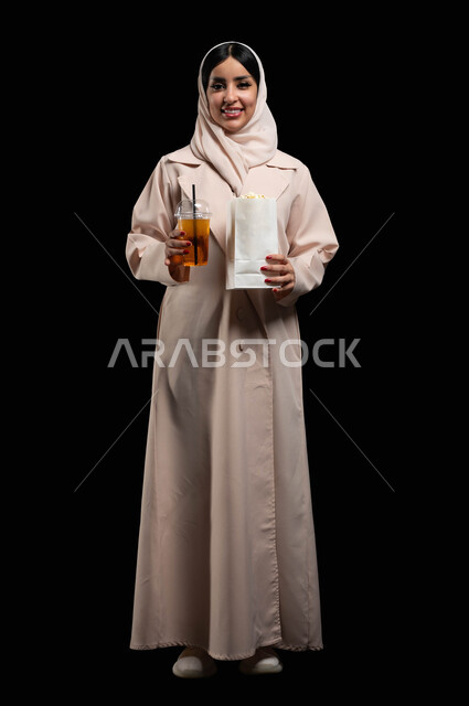 قضاء أوقات ممتعة أثناء العطلة، الاستعداد للذهاب الى السينما لرؤية الفيلم المفضل، بورتريه لامرأة عربية خليجية سعودية محجبة ترتدي عباءه بيج تحمل كيس فوشار وكأس من المشروب الغازي، النظر الى الكاميرا بايماءات الفرح والسعادة، الطول الكامل للجسم، خلفية سوداء