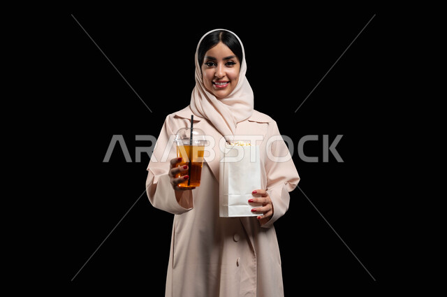 الاستعداد للذهاب الى السينما لرؤية الفيلم المفضل، التسلية والترفيه بقضاء أوقات ممتعة، بورتريه لامرأة عربية خليجية سعودية محجبة ترتدي عباءه بيج تحمل كيس فوشار وكأس من المشروب الغازي، النظر الى الكاميرا بايماءات الفرح والسعادة، خلفية سوداء